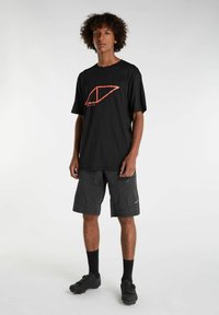 Schwarzes Kurzarmhemd mit rotem geometrischem Design, kombiniert mit einer dunklen Shorts mit Reißverschlusstaschen, getragen mit schwarzen Sportschuhen und Socken.