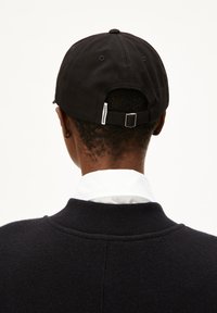 Schwarze Kappe mit gebogenem Schirm, Stofftextur, verstellbarer Riemen hinten, ausgestattet mit einem kleinen Etikett. Getragen mit einem weißen Hemd mit Kragen und einem dunklen Pullover.