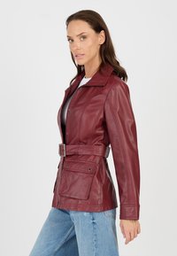 Kvinde iført en tætsiddende bordeaux læderjakke med bælte og store klaplommer, parret med lyseblå jeans mod en hvid baggrund.