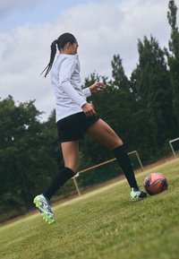 Biely dlhý rukáv, čierne šortky a čierne ponožky po kolená; futbalové kopačky s zelenými akcentmi; Kopanie do viacfarebného futbalového lopty na tráve.