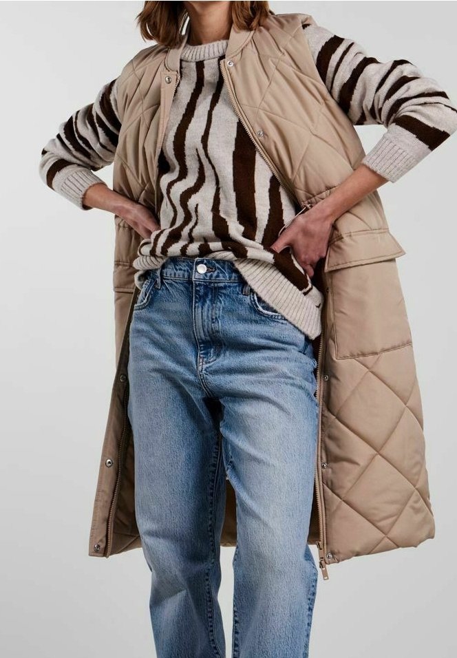 Personne portant un jean bleu clair, un long gilet matelassé beige et un pull beige avec des rayures abstraites marron foncé, debout, les mains sur les hanches.