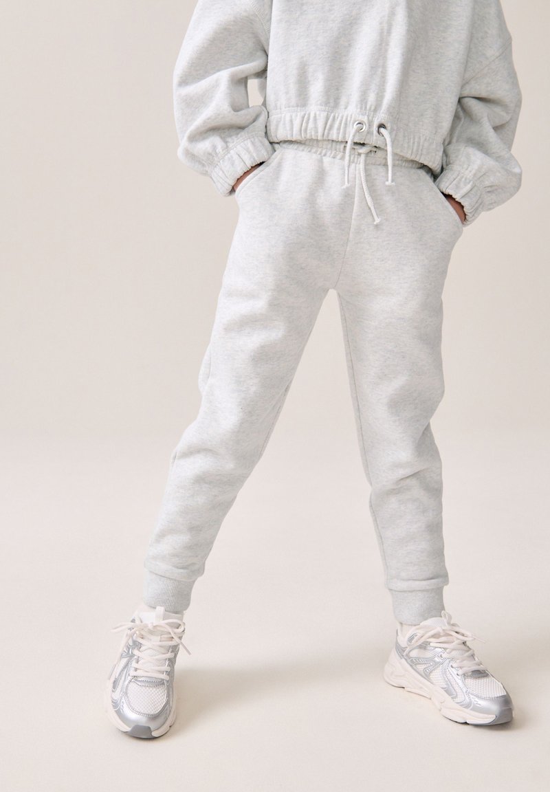 Survêtement gris clair avec des poignets et une taille élastiques, doté de poches. Assorti avec des baskets métalliques argentées et des lacets blancs.