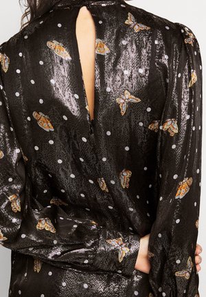 Frau trägt eine glänzende schwarze Bluse mit weißen Punkten und orangefarbenen Schmetterlingsmustern, Rückenschlitz, lange Ärmel und Bündchen an den Handgelenken.