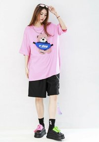 Camiseta rosa de gran tamaño con un gráfico de un oso y pantalones cortos negros. El calzado presenta cordones en colores combinados y calcetines con logotipos. Los accesorios incluyen gafas de sol.