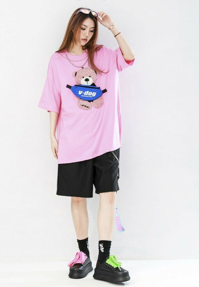 Camiseta rosa de gran tamaño con un gráfico de un oso y pantalones cortos negros. El calzado presenta cordones en colores combinados y calcetines con logotipos. Los accesorios incluyen gafas de sol.