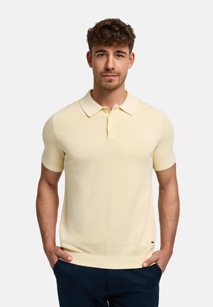 Mand med kort krøllet hår iført en lysegul kortærmet polo og mørke bukser, stående med hænderne i lommerne.