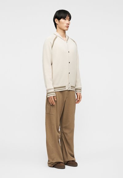 Νεαρός άνδρας όρθιος, φοράει μπεζ ζακέτα με κουμπιά και ριγέ μανσέτες, καθώς και άνετο καφέ παντελόνι με πλαϊνές τσέπες, μπροστά σε απλό φόντο.