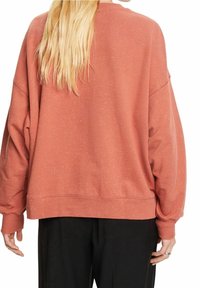 Femme aux longs cheveux blonds portant un sweat-shirt ample en terracotta tacheté et un pantalon noir, vue de dos.