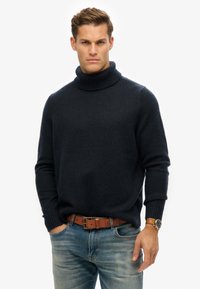 Suéter de cuello alto azul marino en una tela suave y texturizada, con puños y cuello de canalé, combinado con jeans azules deslavados y un cinturón marrón.
