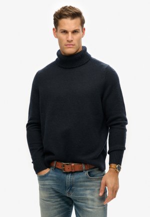 Suéter de cuello alto azul marino en una tela suave y texturizada, con puños y cuello de canalé, combinado con jeans azules deslavados y un cinturón marrón.