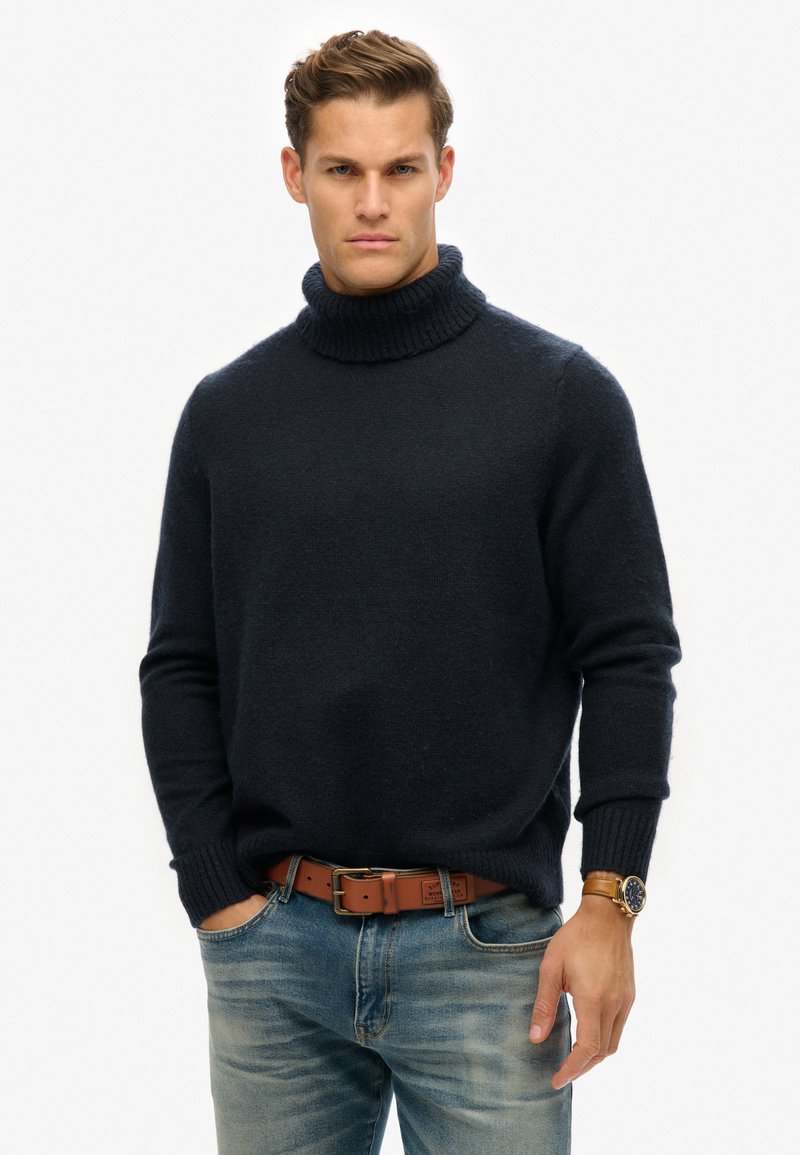 Suéter de cuello alto azul marino en una tela suave y texturizada, con puños y cuello de canalé, combinado con jeans azules deslavados y un cinturón marrón.