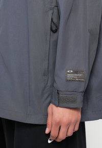 Veste grise en tissu texturé, comportant une poche zippée et un poignet en Velcro. Inclut une étiquette de marque sur la manche.