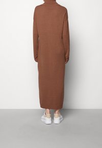 Abito maglione marrone, collo alto a coste, maniche lunghe, lunghezza a metà polpaccio. Abbinato a sneakers bianche con un accento beige sul tallone.