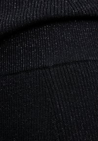 Close-up van zwart ribbelgebreid stof met subtiele metalen draden die een fijne glanseffect op het oppervlak creëren.