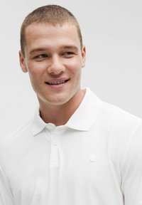 Biele polo tričko s rebrovanou textúrou a zapínaním na dva gombíky. Malé, nenápadné logo je umiestnené na ľavom prsníku.