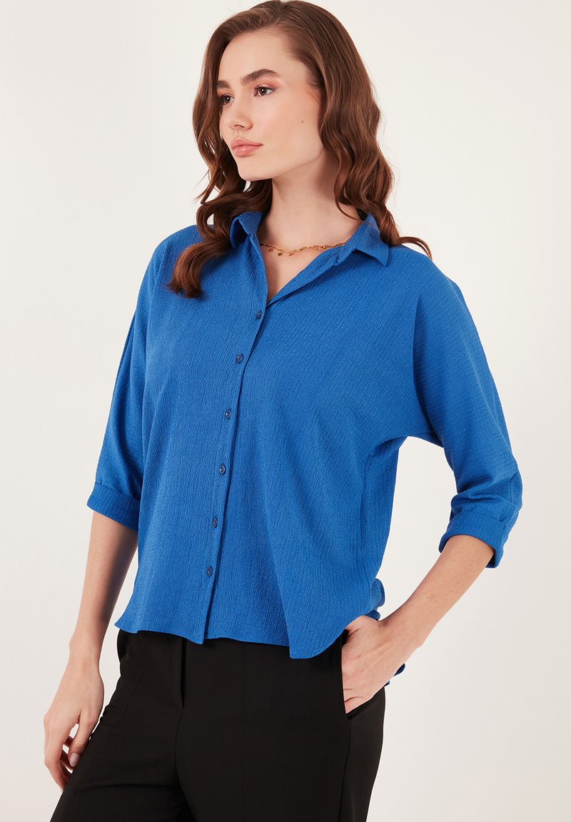 Camisa azul de textura, con botones y mangas remangadas, ajuste relajado y sutiles acentos de botones, combinada con pantalones negros.