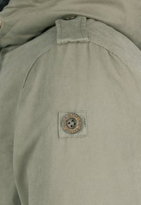 Détail d'une veste kaki avec un bouton rond sur une pièce carrée. Le tissu est lisse avec un éclat subtil et une coupe cintrée.
