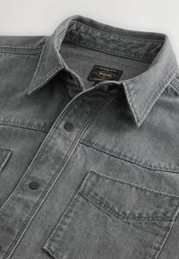 Grå denimskjorte med krave, to forlommer og sorte knapper. Stoffet har en struktureret overflade og synlige syninger.