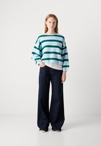 Gestreepte blauwe en groene fuzzy trui met brede mouwen, gecombineerd met losse donkere spijkerbroek. Model staat tegen een effen achtergrond.