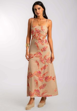 Femme portant une robe maxi sans manches beige avec des motifs de feuilles roses et oranges, un bracelet en or, des colliers superposés et des chaussures compensées beiges.