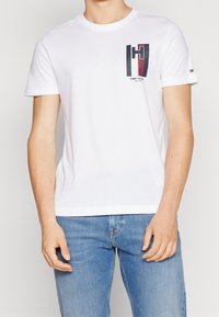 Bílá bavlněná trička s krátkým rukávem, se zeleno-modrým a červeným grafickým logem na levém prsu a malým logem na rukávu.