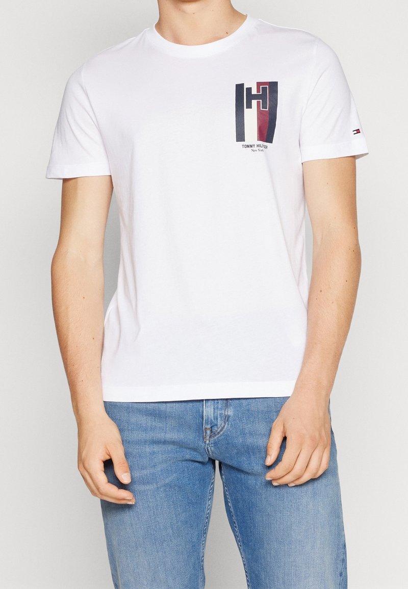 Bílá bavlněná trička s krátkým rukávem, se zeleno-modrým a červeným grafickým logem na levém prsu a malým logem na rukávu.