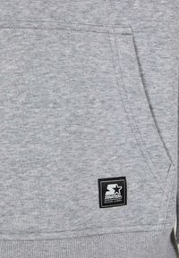 Hanorac gri, texturat, cu buzunar tip kangaroo și tiv cu nervuri. Un patch pătrat negru cu o stea albă și logo-ul "STARTER BLACK LABEL" este vizibil.