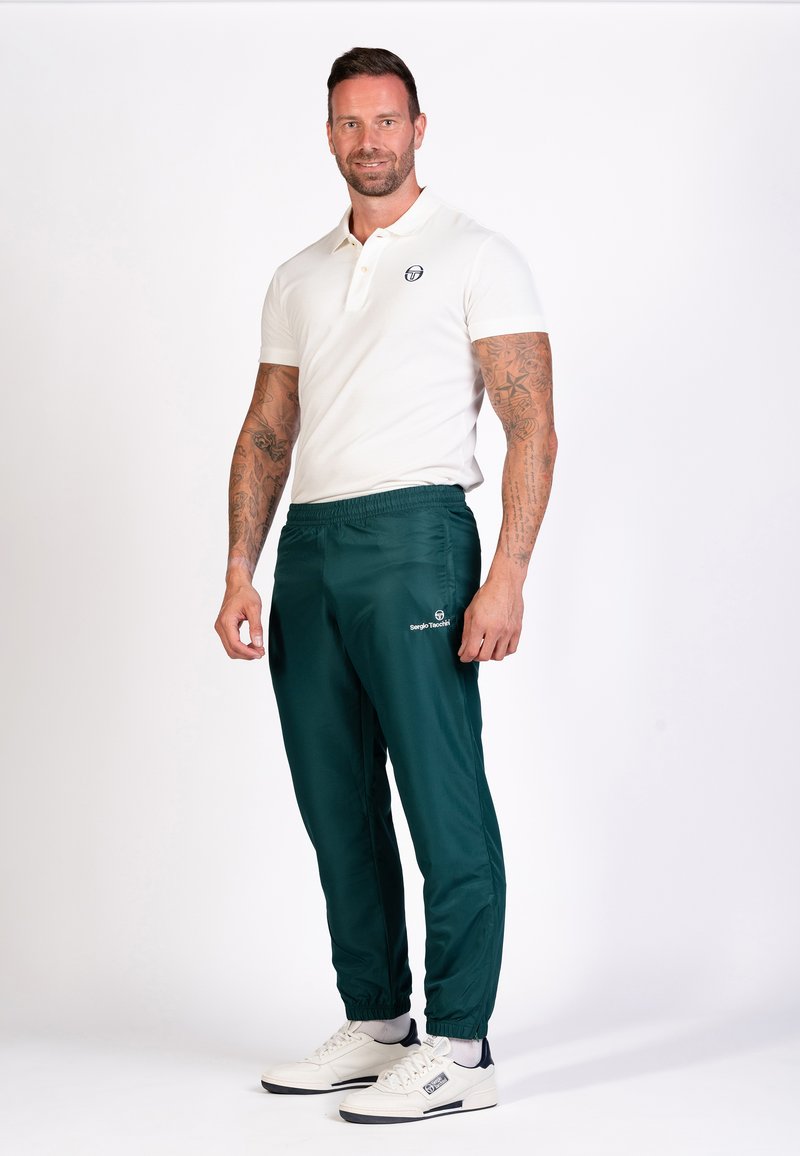 Witte polo met korte mouwen en geborduurd logo, gecombineerd met groene sportbroeken met een elastische tailleband en logo. Witte sneakers.