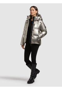 Donna che indossa una giacca a vento argentata metallizzata con cappuccio, pantaloni neri e stivali neri fino al ginocchio, in piedi su uno sfondo semplice grigio chiaro.