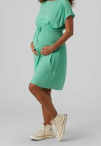Knee-length těhotenské šaty v mátově zelené s plisováním a zavazovacími vazy, spojené s krémovými vysokými teniskami. Jednoduchý a ležérní design.