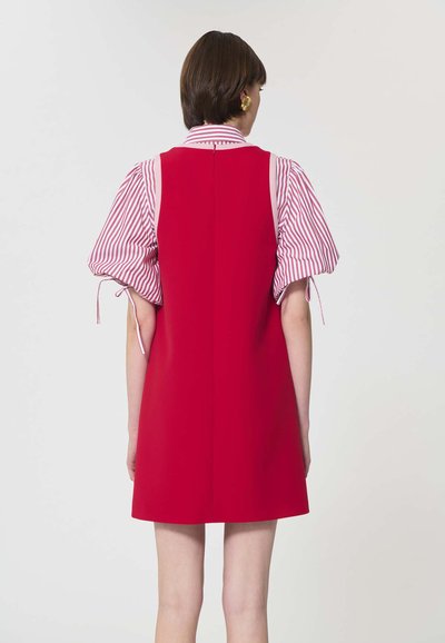 Femme aux cheveux bruns courts portant une robe rouge sans manches sur une chemise à manches bouffantes rayée rose et blanche, debout devant un fond uni clair.