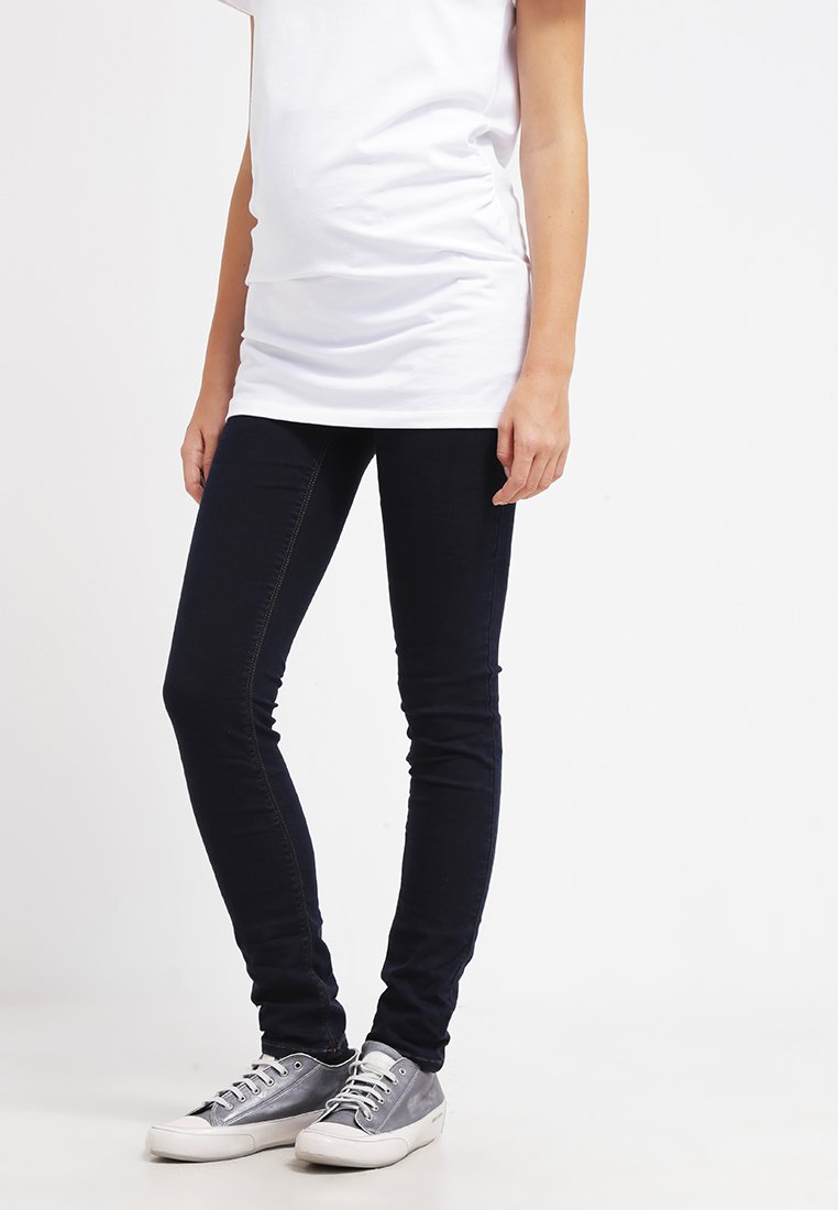 Jeans skinny em denim azul escuro, com uma textura suave e costura subtil, combinados com sapatilhas de tela cinza e uma blusa branca.