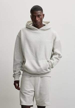 Homme portant un sweat à capuche gris clair et un short assorti, debout avec une main dans la poche devant un fond uni.
