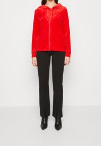 Sudadera con cremallera roja de suave terciopelo con capucha y cordones, combinada con pantalones negros ajustados y botines negros puntiagudos.