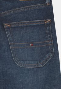 Tmavě modré džínové kalhoty s kapsou a oranžovým prošitím, na pravé straně kapsy s malým logem v červené, bílé a modré barvě.