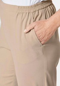 Pantalon beige avec une taille élastique, doté de poches latérales et d'un tissu lisse et léger. Le design comprend une coupe décontractée.