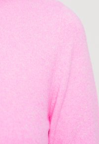 Ροζ πλεκτό πουλόβερ σε κοντινό πλάνο, με ραβδωτή υφή και ελαφρώς χνουδωτή εμφάνιση. Απλός σχεδιασμός χωρίς επιπλέον μοτίβα ή διακοσμητικά στοιχεία.