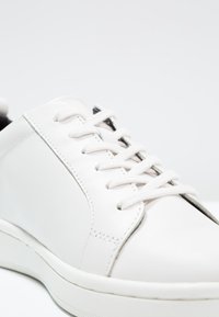 Gros plan d'une basket en cuir blanche avec des lacets blancs et une semelle en caoutchouc texturée sur un fond clair uni.