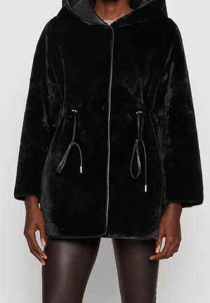 Schwarze Kapuzenjacke aus Kunstfell mit Frontreißverschluss und verstellbaren Kordeln, getragen zu dunkler Hose an einer stehenden Person.