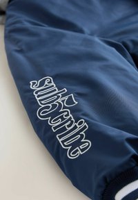 Ärmel einer dunkelblauen Jacke mit weiß gesticktem Text "subscribe", mit einer glatten, glänzenden Oberfläche und kontrastierendem geripptem Bündchen.