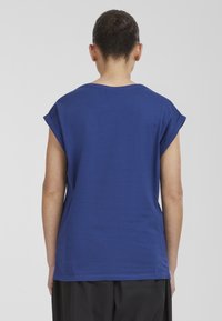 Persoon met donker haar in een bun, gekleed in een effen blauwe korte mouw shirt en zwarte broek, staande tegen een effen lichtgrijze achtergrond.