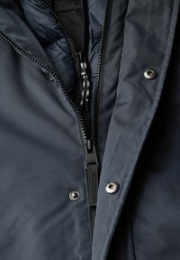 Chaqueta azul oscuro con un exterior suave y texturizado, que presenta un zipper grueso, botones a presión y un forro acolchado interior para aislamiento.