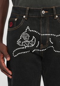 Sorte denimjeans med et krystallbesatt kattedesign, kontrasterende oransjefargede sømmer, og en rød aksent på frontlommen.