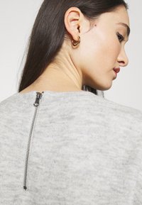 Maglione grigio con una texture sottile, scollatura rotonda e una breve chiusura con zip sul retro. Orecchino a cerchio dorato visibile sul modello.