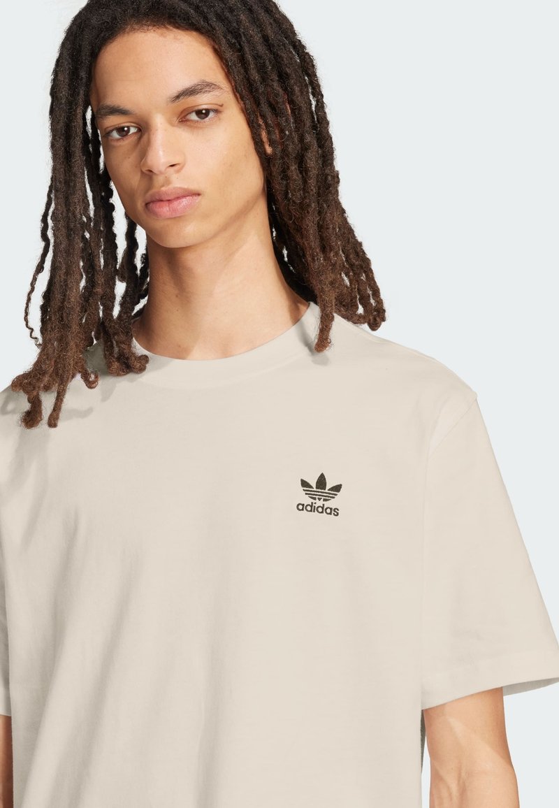 Joven con rastas largas, vistiendo una camiseta beige de Adidas con logo negro en el pecho, fondo neutro.