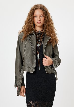 Krótkia, oliwkowa skórzana kurtka o zniszczonej teksturze, z zamykanymi na zamek kieszeniami i srebrnymi detalami, zestawiona z czarną koronkową sukienką.