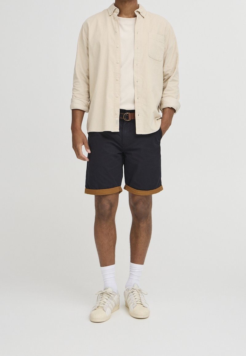 Chemise beige clair boutonnée portée sur un t-shirt crème, associée à un short bleu marine avec des bords marron et des baskets de couleur claire.