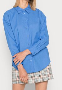 Camicia button-up di colore azzurro chiaro con colletto, maniche lunghe e una texture sottile, abbinata a una gonna a scacchi nei colori pastello.