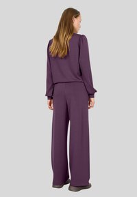Violet loungewear komplektas, kuriame yra puloverio tipo viršus su pūstomis rankovėmis ir plačiomis kelnėmis. Minkšta tekstūra, laisvas siluetas ir minimalus dizainas.