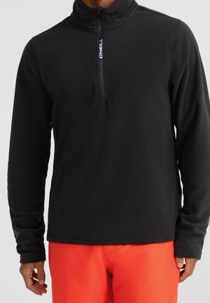 Mann trägt schwarzen Fleecepulli mit langem Arm und Viertel-Reißverschluss und leuchtend orangefarbene Hose vor schlichtem hellem Hintergrund.